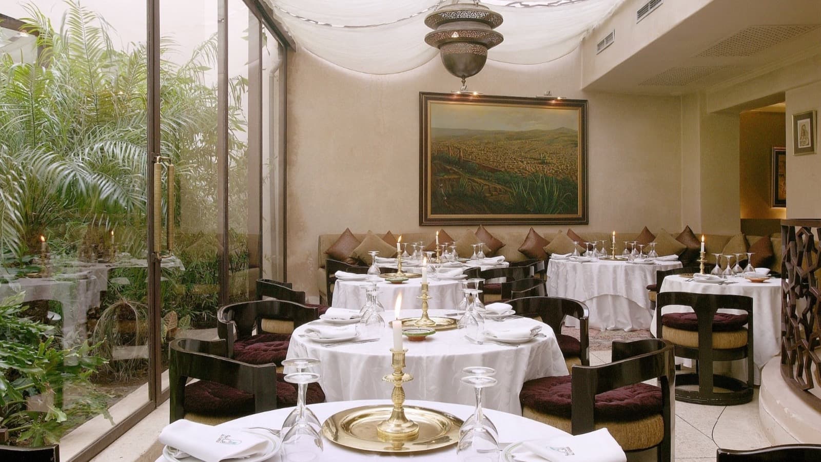 La salle d'Al Fassia Guéliz, tables nappées de blanc, chandeliers de laiton et patio planté derrière une verrière