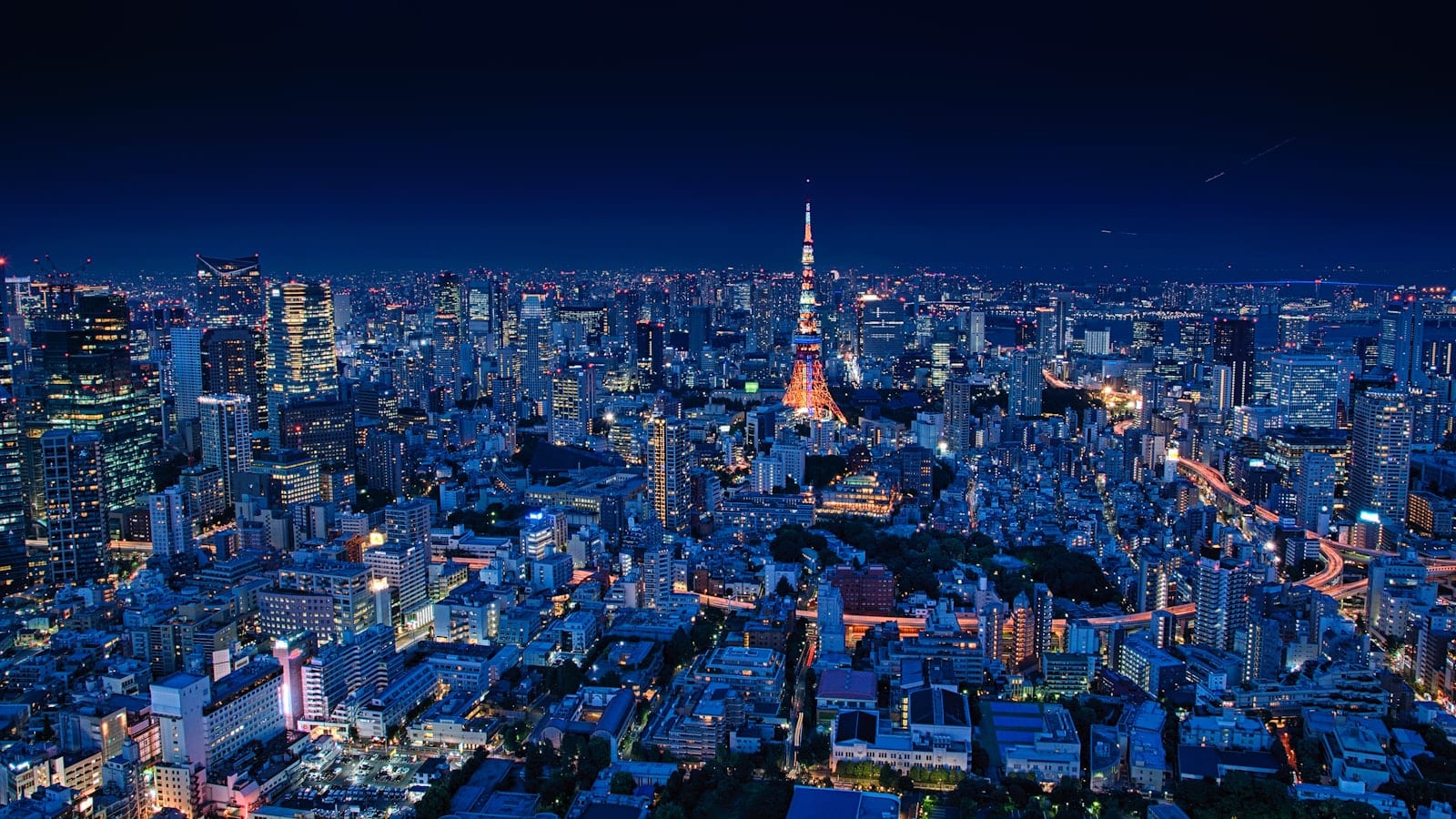 Vue plongeante sur un carrefour de Tokyo à la tombée du jour, avec les tours de bureaux éclairées en arrière-plan et les rames d'une ligne ferroviaire surélevée au premier plan.